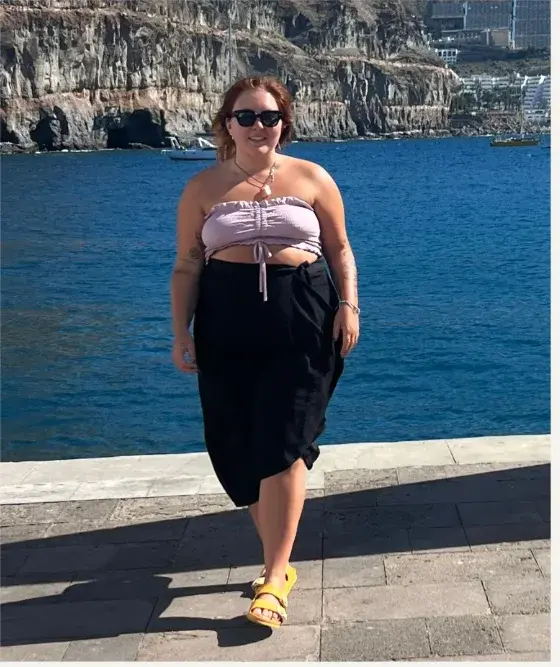 Mathilda kävelee rannalla mustassa hameessa ja vaaleanpunaisessa toppi, aurinkolasit päässä.